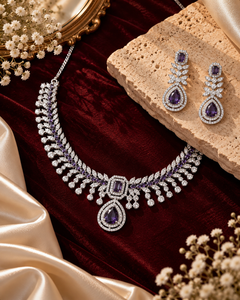 A luxury jewelry set featuring a silver-toned necklace and matching drop earrings with deep purple teardrop gemstones and clear pavé accents, styled on a dark red velvet background with beige silk, a stone slab, and dried flowers.