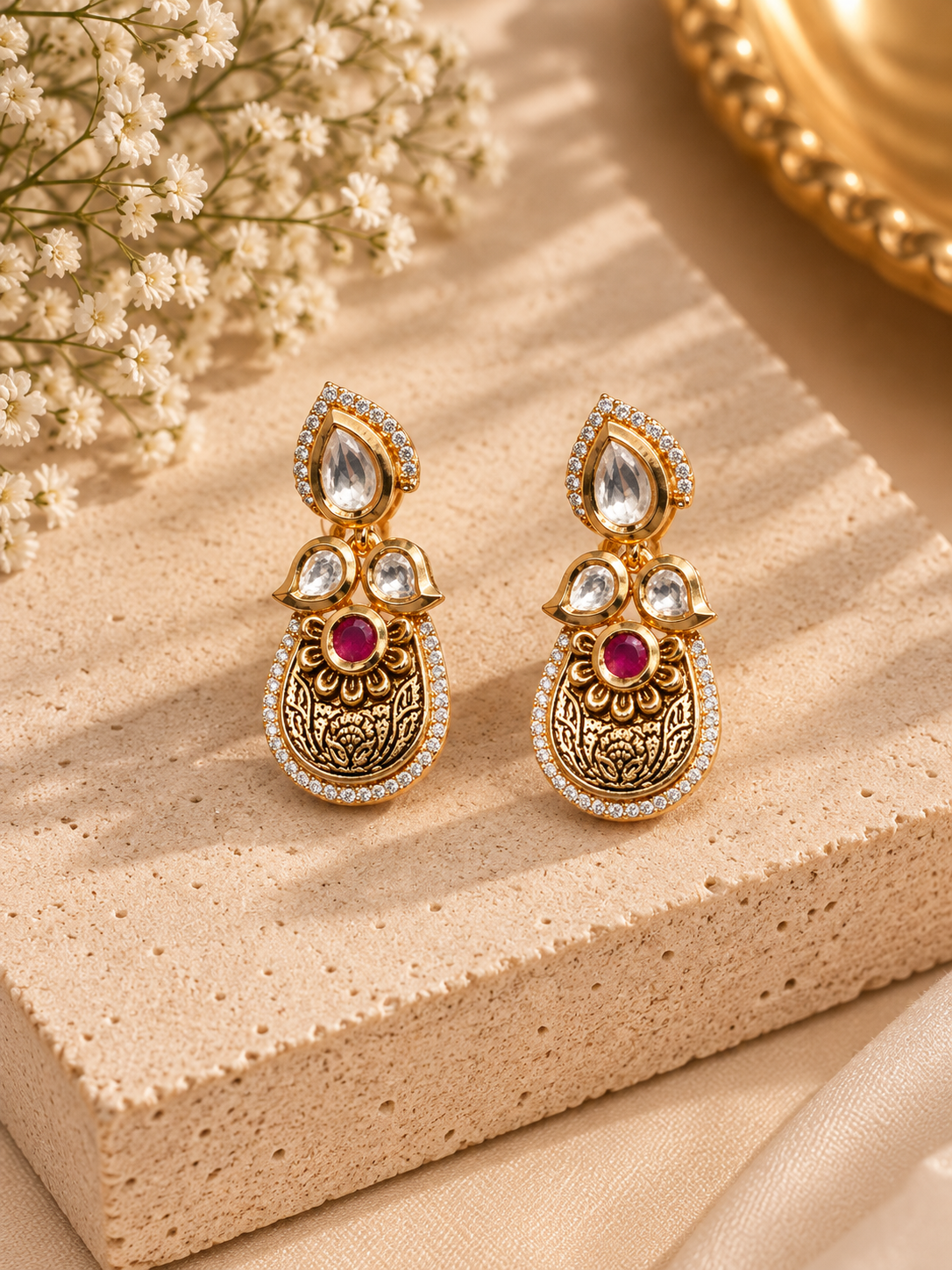 A pair of ornate gold-toned Indian-style earrings with clear stones and red center stones, displayed on a beige stone block with dried flowers in the background.
